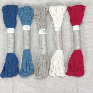 May include: Five skeins of Sashiko thread in various colors: deep blue, light blue, gray, white, and red. Each skein is wrapped with a paper label featuring Japanese text and the brand name "SASHIKO". The threads are neatly arranged on a neutral-toned surface.