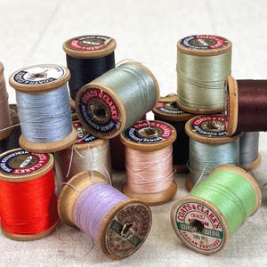 May include: A collection of vintage wooden spools of Coats & Clark's sewing thread in various colors including red, light blue, lavender, green, brown, and pink. Each spool features a circular paper label on the top with brand text and details.