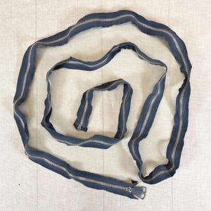 May include: A long, continuous dark navy blue zipper tape arranged in a loose, spiraling coil. The zipper features metallic silver teeth and a silver metal slider pull at one end, with the fabric tape forming a flexible, winding shape.