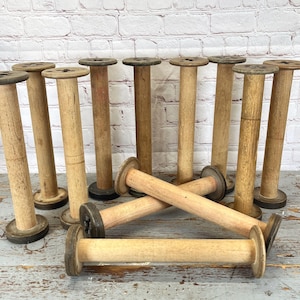 May include: A collection of vintage wooden spools in various sizes. The spools are made of light-colored wood with dark tops and bases. Some spools are standing upright, while others are lying down. The spools are arranged on a weathered wooden surface.