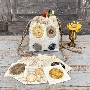 May include: A beige patchwork drawstring bag with a braided cord strap, filled with colourful threads and pens. The bag features embroidered circular designs in various colours. A small brass vase with yellow flowers and pins sits nearby.
