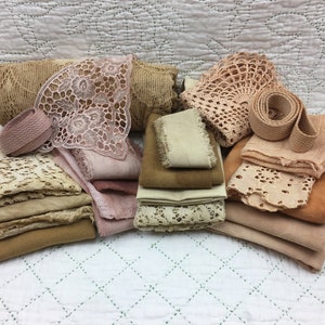 May include: A collection of vintage lace, fabric scraps, and trim in shades of brown and pink. The fabrics are arranged in a pile on a white quilted surface.