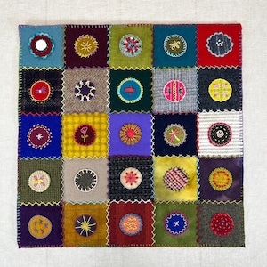 May include: A square patchwork textile featuring a grid of colorful squares. Each square is a different color and texture, with embroidered circular designs in the center. The color palette includes blues, greens, reds, and yellows, with white stitching.
