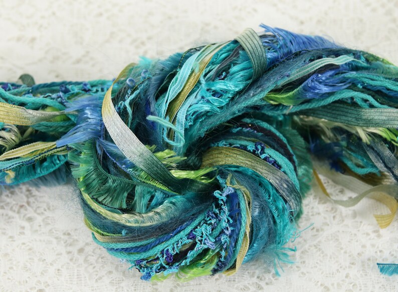 May include: A skein of colorful yarn with a mix of textures, including frayed edges and smooth ribbons. The yarn is in shades of blue, green, and gold.