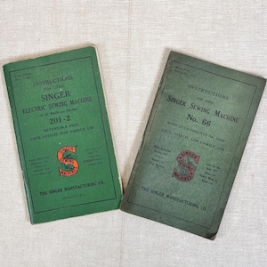 May include: Two vintage green paper instruction manuals for Singer sewing machines. The left manual is for the 201-2 model, and the right manual is for the No. 66 model. Both feature black text, the Singer logo, and The Singer Manufacturing Co. name.