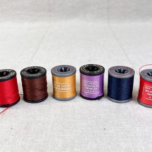 May include: Six small cylindrical spools of sewing thread in red, brown, gold, purple, navy blue, and bright red. Each spool features a black or grey plastic top and bottom with the brand name Molnlycke printed on the top edge of the spool.