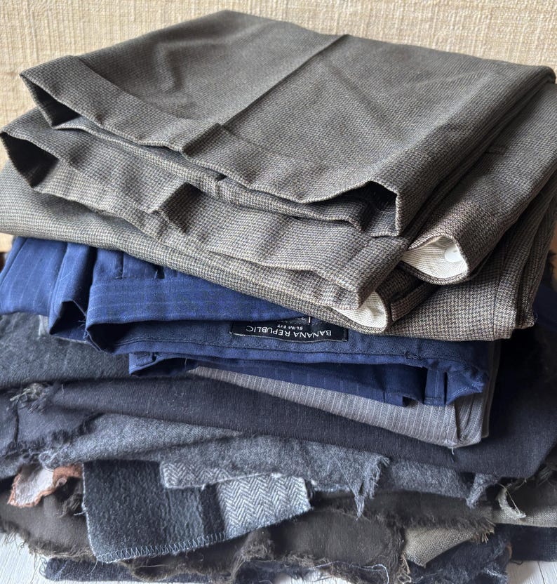 May include: A stack of folded trousers in various shades of gray, blue, and black. The top layers are tailored pants with cuffs, while the lower layers appear to be fabric swatches. One pair of pants has a "Banana Republic" label.