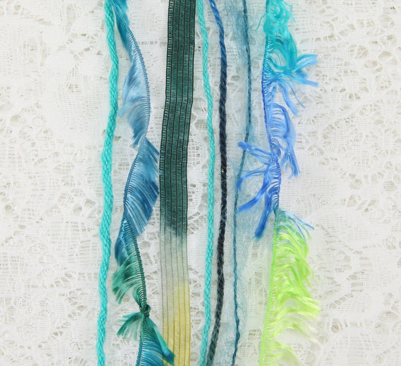 May include: Assortment of colorful yarn and ribbon strands with fringed edges, hanging against a white lace background.