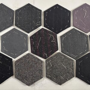May include: A collection of hexagonal fabric patches in various shades of gray, black, and purple. Each patch is stitched with white thread around the edges. The fabrics have different textures, including pinstripes and tweed.