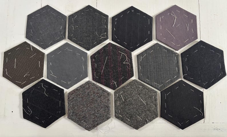 May include: A collection of hexagonal fabric patches in various shades of gray, black, and purple. Each patch is stitched with white thread around the edges. The fabrics have different textures, including pinstripes and tweed.