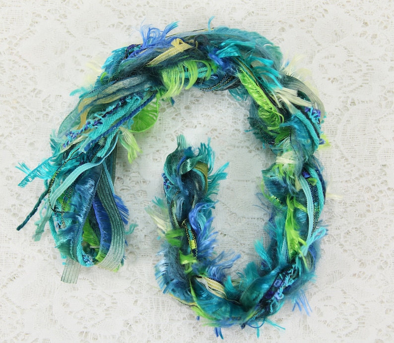 May include: A bundle of colorful yarn in shades of blue, green, and white. The yarn is made of a variety of textures, including fuzzy, silky, and ribbon-like strands.
