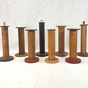 May include: A collection of eight vintage wooden spools in various sizes and finishes. The spools have cylindrical bodies and circular bases, with some featuring decorative tops. Colors include natural wood tones, red, and black.