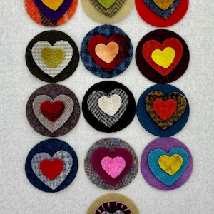 May include: A collection of 14 felt circles with layered hearts in various colors and patterns. The hearts are in shades of red, pink, yellow, green, and white. The circles are in shades of brown, blue, purple, and black.
