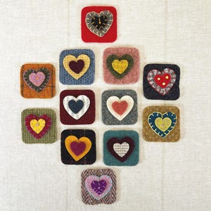 May include: A collection of twelve square, felted coasters, each featuring a heart design. The hearts are in various colors, including red, yellow, blue, and pink, with contrasting stitching. The coasters are arranged on a neutral background.