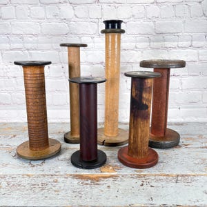 May include: A collection of six vintage wooden spools in various sizes and finishes. The spools feature a range of colors, including light and dark browns, and a dark burgundy. Some have a textured appearance, while others are smooth.