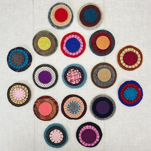 May include: A collection of colorful, circular felt coasters. Each coaster features concentric circles of various colors, including red, blue, pink, and green. The coasters are arranged on a neutral-colored surface, showcasing their vibrant designs.