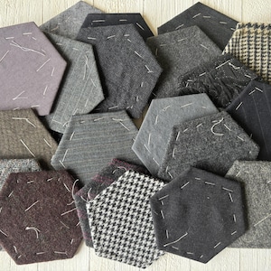 May include: A collection of hexagonal fabric coasters in various shades of gray, brown, and black. Each coaster features a unique pattern, including herringbone, pinstripe, and solid textures, with white stitching details. The coasters are arranged in a visually appealing, overlapping pattern.