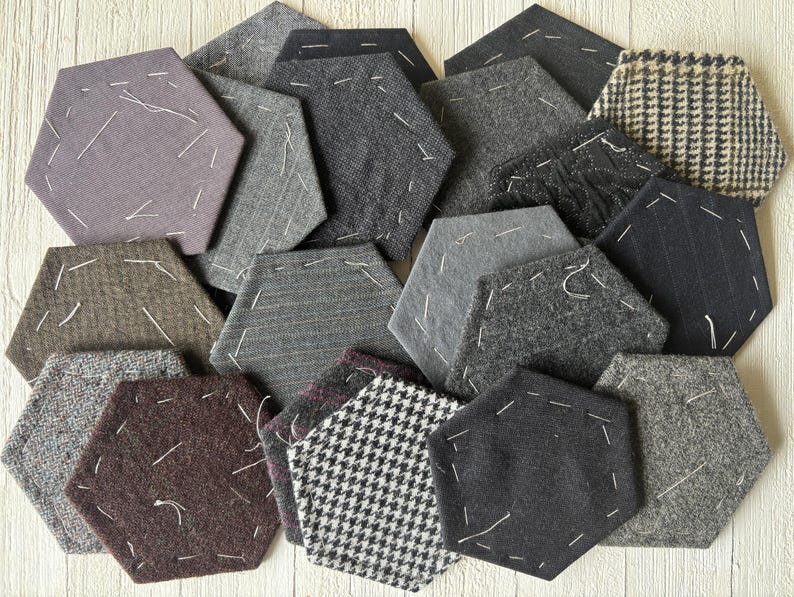 May include: A collection of hexagonal fabric coasters in various shades of gray, brown, and black. Each coaster features a unique pattern, including herringbone, pinstripe, and solid textures, with white stitching details. The coasters are arranged in a visually appealing, overlapping pattern.