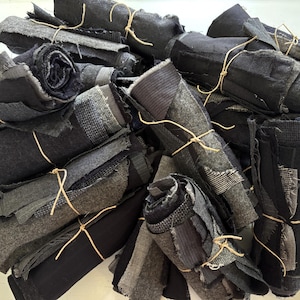 May include: A pile of rolled fabric scraps in various shades of gray and black, tied with twine. The fabric pieces have different textures and patterns, including stripes and woven designs. The overall aesthetic is rustic and industrial.