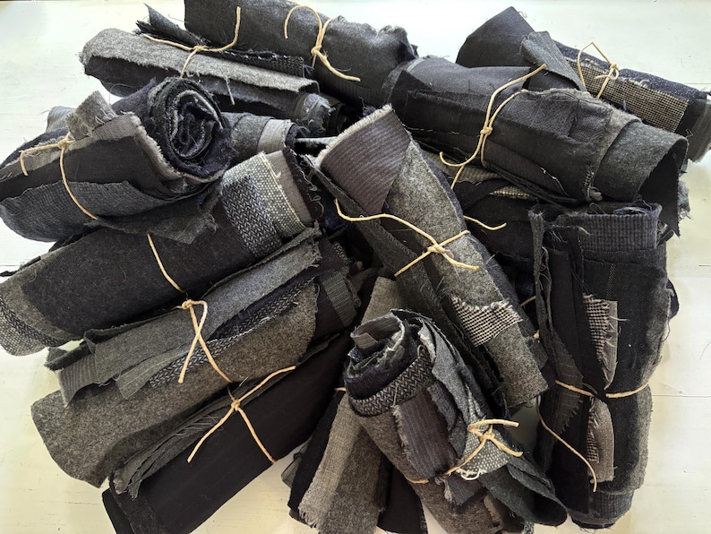 May include: A pile of rolled fabric scraps in various shades of gray and black, tied with twine. The fabric pieces have different textures and patterns, including stripes and woven designs. The overall aesthetic is rustic and industrial.