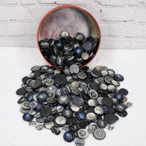 May include: A large pile of black, gray, and blue plastic buttons in a variety of sizes and styles. The buttons are spilling out of a red tin container.