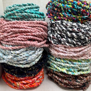 May include: A collection of coiled fabric ropes in various colors and patterns, including teal, dusty pink, light blue, navy, orange, multicolored, black and white, and brown. Each rope is constructed from braided textile strips.