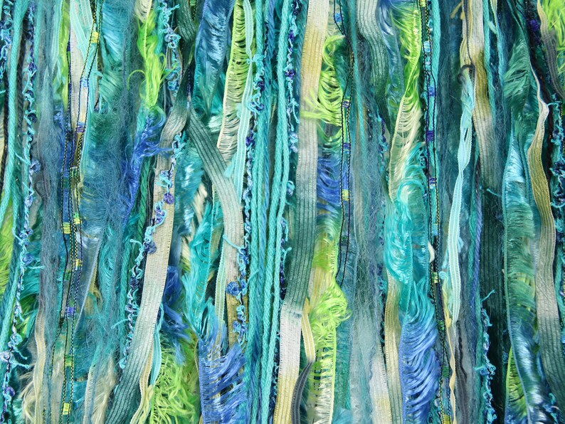 May include: A close-up of a wall hanging made of various shades of blue, green, and white yarn. The yarn is woven together to create a textured and colorful design.