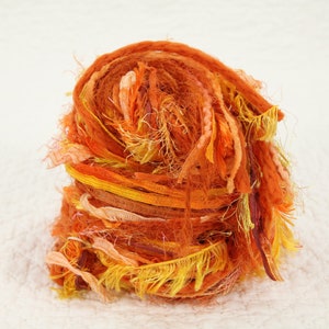 May include: A ball of orange, yellow, and peach colored yarn made from recycled fabric scraps. The yarn is loosely wound and has a fluffy texture.