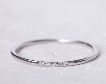 May include: A delicate silver ring with five small, clear gemstones set in a row. The ring is thin and has a polished, reflective surface, resting on a white, textured background. The ring is a piece of jewelry.