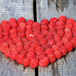 May include: A heart shape made of bright red raspberries on a weathered gray wooden surface. The raspberries are in focus, showcasing their textured surface and vibrant color. The arrangement is centered, creating a visually appealing composition.