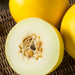 May include: A yellow cantaloupe melon cut in half, revealing the white flesh and brown seeds. The melon is sitting on a woven wicker surface.