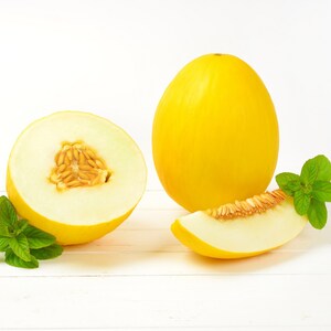 May include: A yellow cantaloupe melon cut in half with a slice of melon and green mint leaves on a white surface.