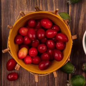 May include: Overhead shot of a wooden table with three bowls. A yellow wooden bowl is filled with bright red berries. A wooden bowl on the left contains hazelnuts. A white bowl on the right holds green feijoa fruits.