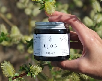 May include: A black glass jar with a silver lid and a white label that says "LJÖS FREMJJA". The jar is being held by a hand in front of a background of green foliage with yellow flowers.