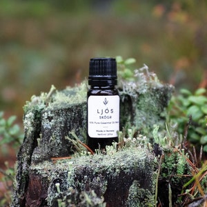 Peut inclure: Un petit flacon en verre brun foncé avec un bouchon noir, étiqueté "LJÖS SKOGR 100% Pure Essential Oil", posé sur une souche d'arbre recouverte de mousse dans un environnement forestier.