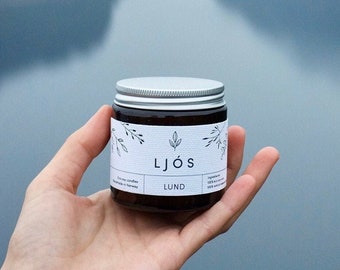 May include: A brown glass candle jar with a silver lid. The label is white with black text that reads "LJÖS" and "LUND". The candle is being held in a hand against a blurry background of a cloudy sky.