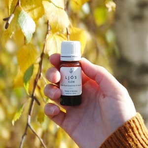 May include: A small brown glass bottle with a white label that says "LJOS ELDR 100% Pure Essential Oil Blend Made in Norway Net volume: 10 ml". The bottle is being held in a hand wearing a brown sweater against a background of yellow and green leaves.