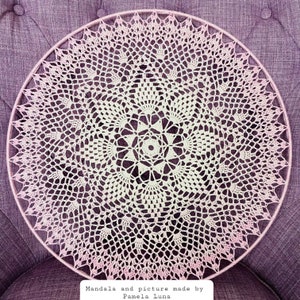 Puede incluir: Un mandala blanco tejido a crochet con un borde rosa degradado, colgado sobre un fondo de tela morado. El texto "Mandala and picture made by Pamela Luna" est&aacute; en la parte inferior de la imagen.