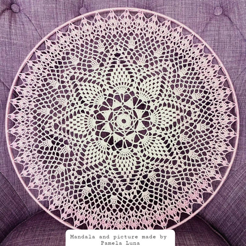 Puede incluir: Un mandala blanco tejido a crochet con un borde rosa degradado, colgado sobre un fondo de tela morado. El texto "Mandala and picture made by Pamela Luna" est&aacute; en la parte inferior de la imagen.