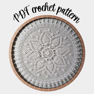 Puede incluir: Un encaje de ganchillo blanco en un aro de bordado de madera. El encaje tiene un dise&ntilde;o delicado e intrincado con un motivo floral central. El texto "PDF crochet pattern" est&aacute; escrito encima del aro.