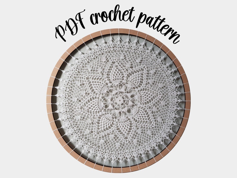 Puede incluir: Un encaje de ganchillo blanco en un aro de bordado de madera. El encaje tiene un dise&ntilde;o delicado e intrincado con un motivo floral central. El texto "PDF crochet pattern" est&aacute; escrito encima del aro.