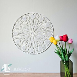 Op de afbeelding: Een wit gehaakt doily hangt aan een witte muur met een boeket gele, roze en witte tulpen in een donkergrijze vaas op een houten tafel.