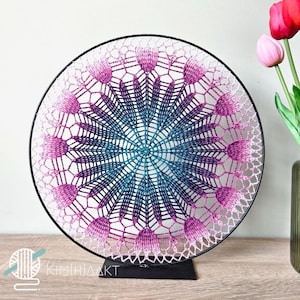 Op de afbeelding: Een gehaakt mandala met een gradiënt van roze, paarse en blauwe kleuren wordt getoond op een zwarte metalen standaard. De mandala is rond en heeft een delicaat, ingewikkeld ontwerp.