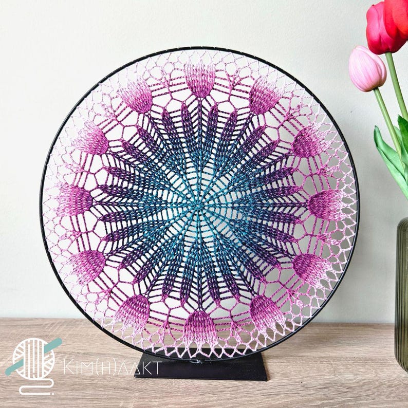 Op de afbeelding: Een gehaakt mandala met een gradi&euml;nt van roze, paarse en blauwe kleuren wordt getoond op een zwarte metalen standaard. De mandala is rond en heeft een delicaat, ingewikkeld ontwerp.