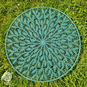 May include: A teal circular crocheted doily or placemat. The intricate openwork design features a repeating leaf-like pattern radiating from the centre. The doily is placed on green grass, creating a contrast of textures and colours.