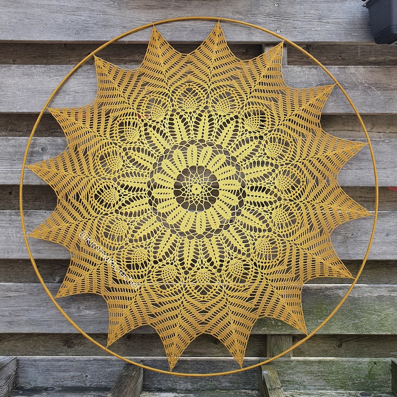 Puede incluir: Un gran doily de ganchillo amarillo con un dise&ntilde;o de estrella est&aacute; expuesto en una pared de madera. El doily est&aacute; enmarcado por un aro de metal circular.