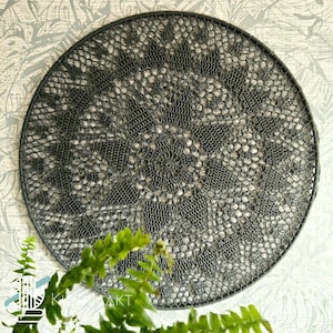 May include: A circular dark gray crochet wall hanging featuring an intricate, symmetrical lace pattern with a central starburst design. The delicate, openwork textile is stretched tightly across a matching dark gray circular frame.