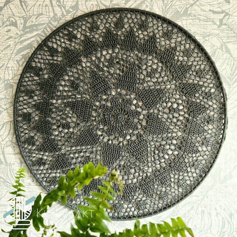 May include: A circular dark gray crochet wall hanging featuring an intricate, symmetrical lace pattern with a central starburst design. The delicate, openwork textile is stretched tightly across a matching dark gray circular frame.