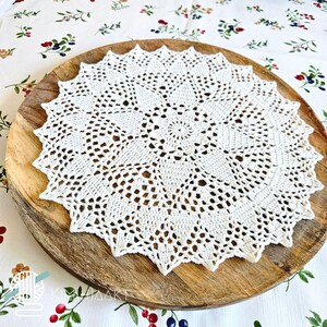 May include: A round, off-white crochet doily featuring an intricate, star-shaped geometric pattern with delicate openwork stitching. The circular textile piece displays a central floral-like motif that radiates outward into pointed, scalloped edges.