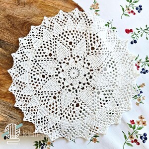 May include: A round, off-white crochet doily featuring an intricate, radiating starburst pattern with openwork stitches and a scalloped edge. The design consists of a central circular motif surrounded by repeating geometric segments.